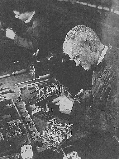 Ein älterer Feinmechaniker mit Brille montiert ein Taxameterwerk, im Hintergrund weitere Arbeiter.