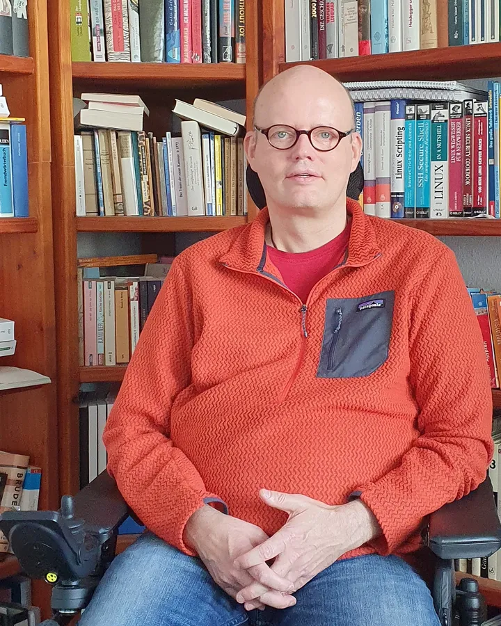 Peter Haufschild sitzt im Rollstuhl vor einem vollen Bücherregal; er trägt eine runde Brille und einen roten Pullover.